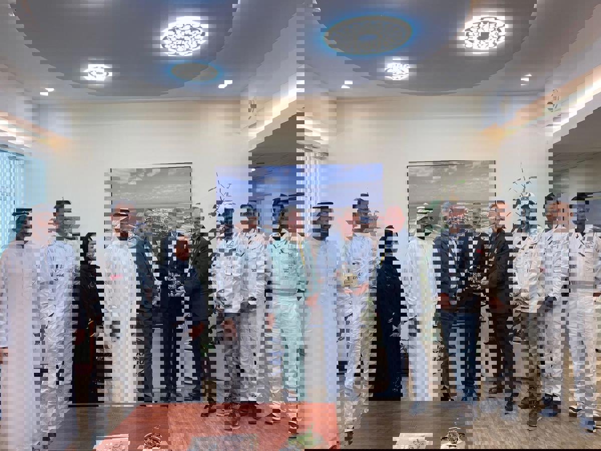 Group photo taken indoors during a visit to Drydocks World Dubai. Several people are standing in a meeting room, some wearing white coveralls with company logos and others in formal business attire. A person in a green suit is positioned near the center, and a small trophy or award is being presented. Behind the group is a large wall-mounted screen displaying an aerial view of a port facility. The room has modern lighting, plants, and a wooden table in the foreground.