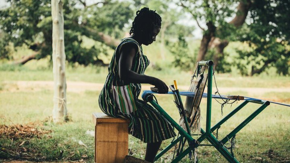 A female textile worker in Ghana uses a manual hand machine to weave cloth
