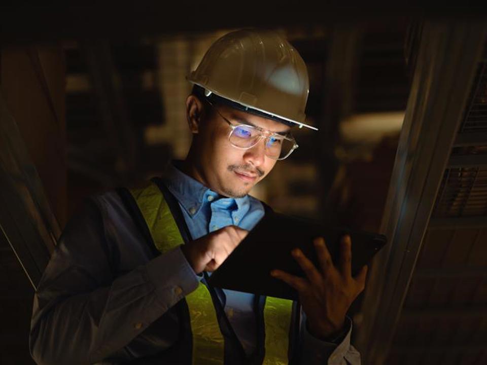 A person wearing a hard hat and reflective safety vest stands in a dim industrial or construction setting, using a tablet device held in one hand while tapping the screen with the other.
