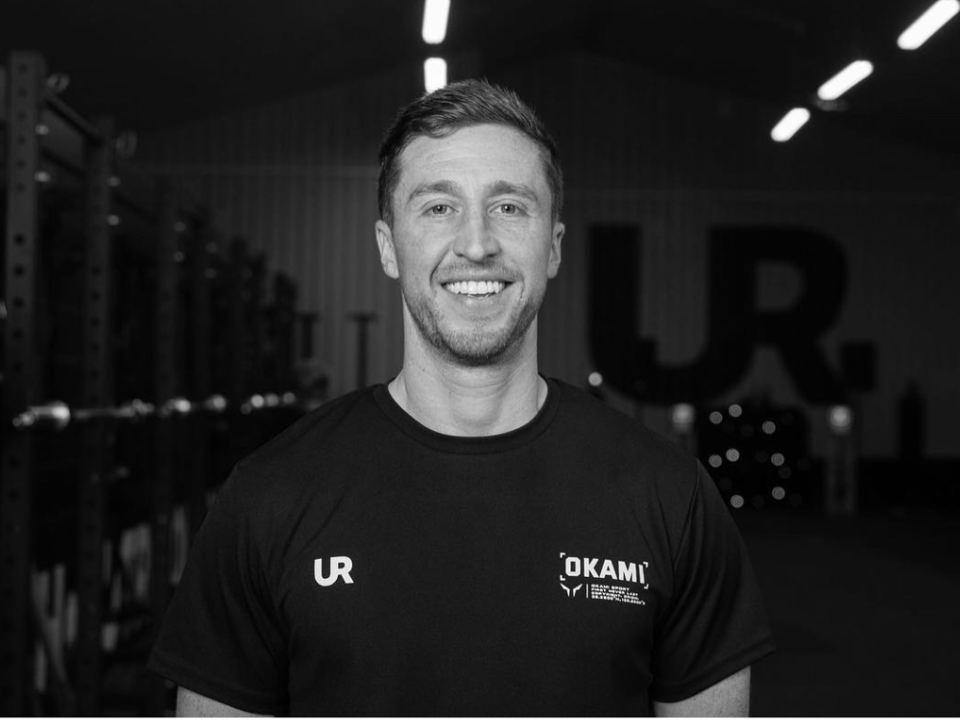 A person in a black “Urban Reform” hoodie works on a laptop at a desk near large windows.