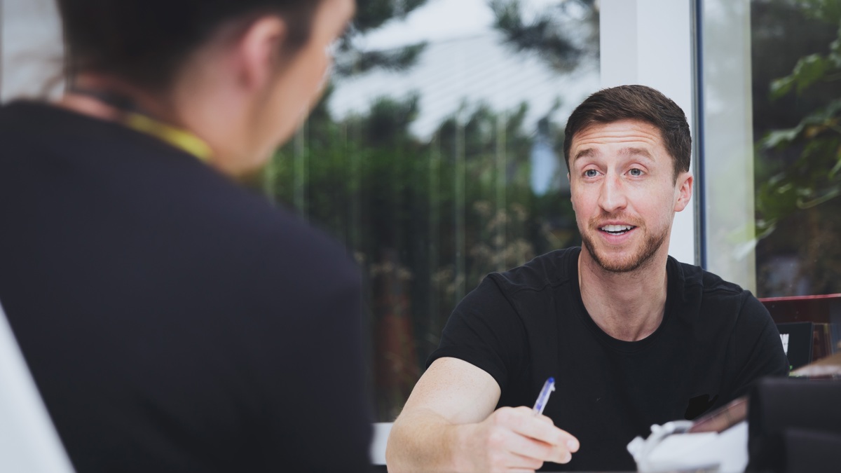 A person sits at a table writing while speaking with another individual across from them in a bright, indoor setting.