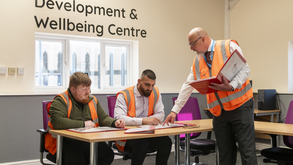 Worker in high-visibility vest reviewing documents with colleagues in a training room labelled “Development & Wellbeing Centre”