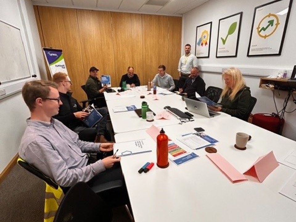 People seated around a long white conference table during a training session. Laptops, notebooks, and printed materials are on the table. A person stands at the back leading the session. Posters with sustainability graphics and a whiteboard are visible.